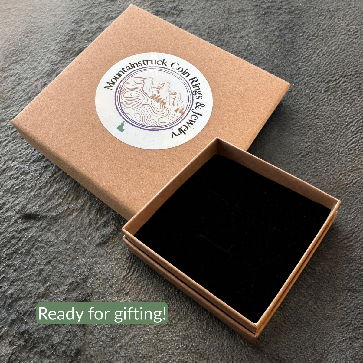 A brown kraft gift box with a round white sticker reading Mountaintruck Coin Jewelry, shown closed with a green diagonal 'Ready for gifting!' text overlay and next to an open box containing a black velvet insert, set on a gray stone surface. 
- Mountainstruck Coin Jewelry
