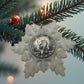 2025 Half Dollar Christmas Ornament   (glows) with an uncirculated half dollar cased inside a white 3D printed snowflake with golden tassel on a pine tree background. - Mountainstruck Coin Jewelry