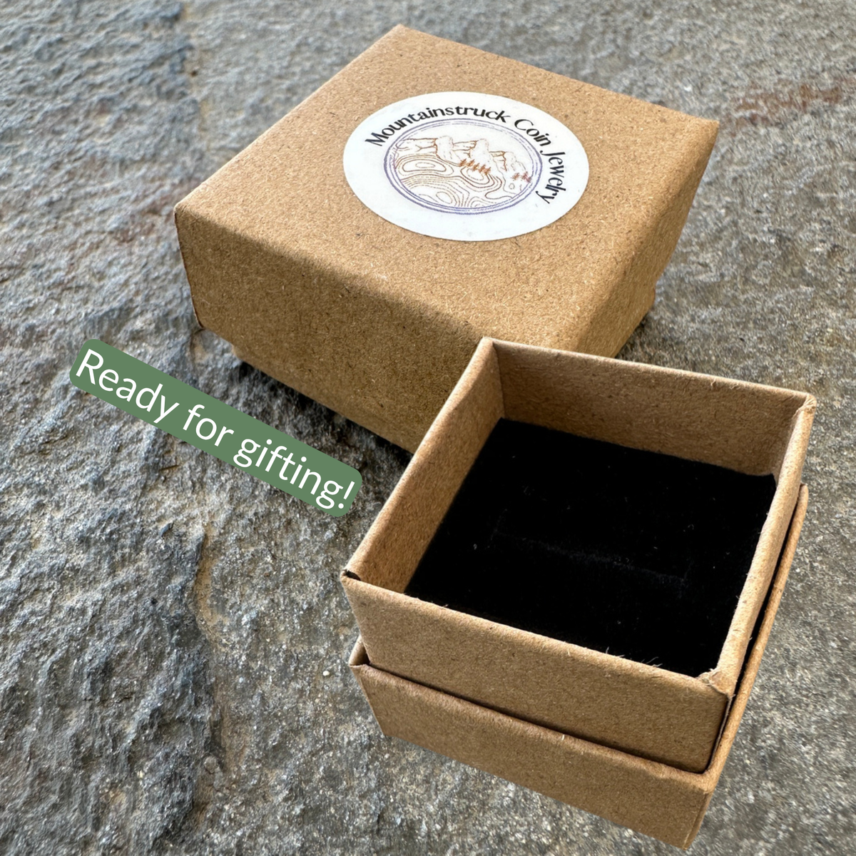 A brown kraft gift box with a round white sticker reading Mountaintruck Coin Jewelry, shown closed with a green diagonal 'Ready for gifting!' text overlay and next to an open box containing a black velvet insert, set on a gray stone surface. 
- Mountainstruck Coin Jewelry