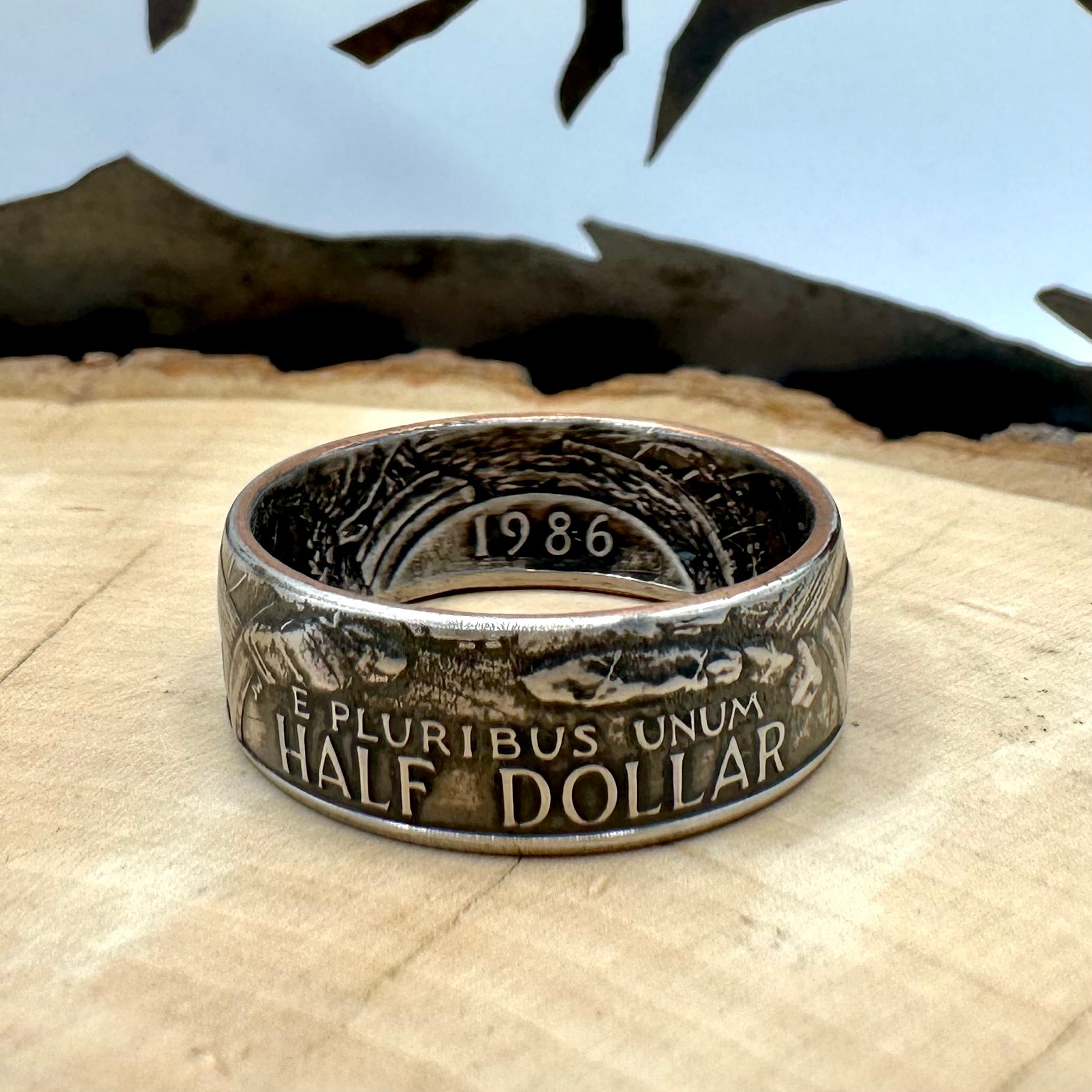 "A Nation of Immigrants" Half Dollar Coin Ring patina on light wood round with a metal mountain background. - Mountainstruck Coin Jewelry