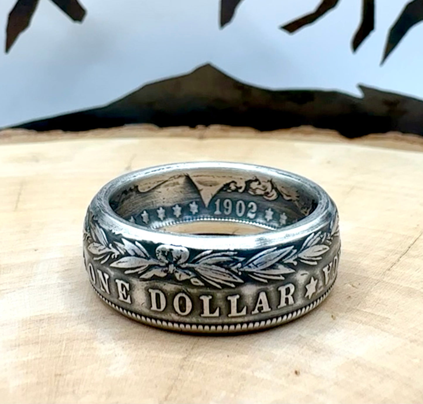 Morgan Dollar Silver Coin Ring patina on a light wood round with a metal mountain background. - Mountainstruck Coin Jewelry