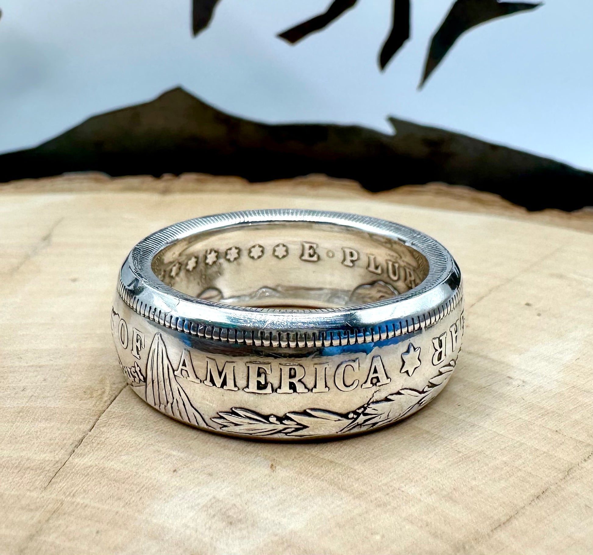 Morgan Dollar Silver Coin Ring shine on a light wood round with a metal mountain background. - Mountainstruck Coin Jewelry