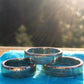 Idaho, Maine, California U.S. State Quarter Coin Rings shined stacked on a blue cloth with sun streaming in front of a blurred green pine tree background. - Mountainstruck Coin Jewelry