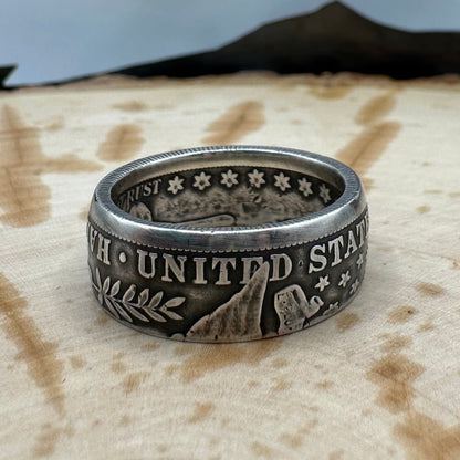 U.S. Barber Half Dollar Silver Coin Ring (reverse) on a light wood round with a metal mountain background. - Mountainstruck Coin Jewelry