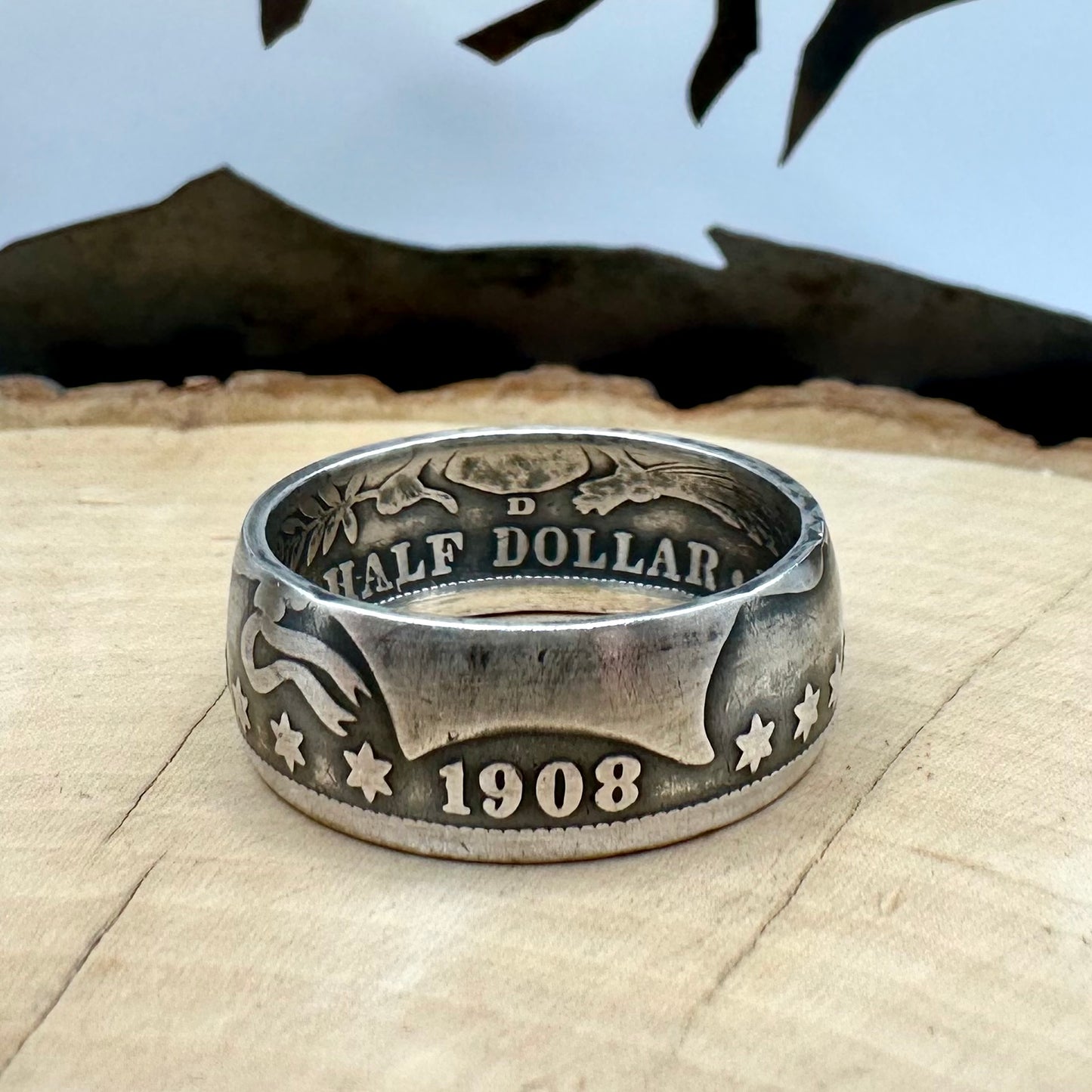 U.S. Barber Silver Half Dollar Coin Ring (front) on a light wood round with metal mountains in the background. - Mountainstruck Coin Jewelry