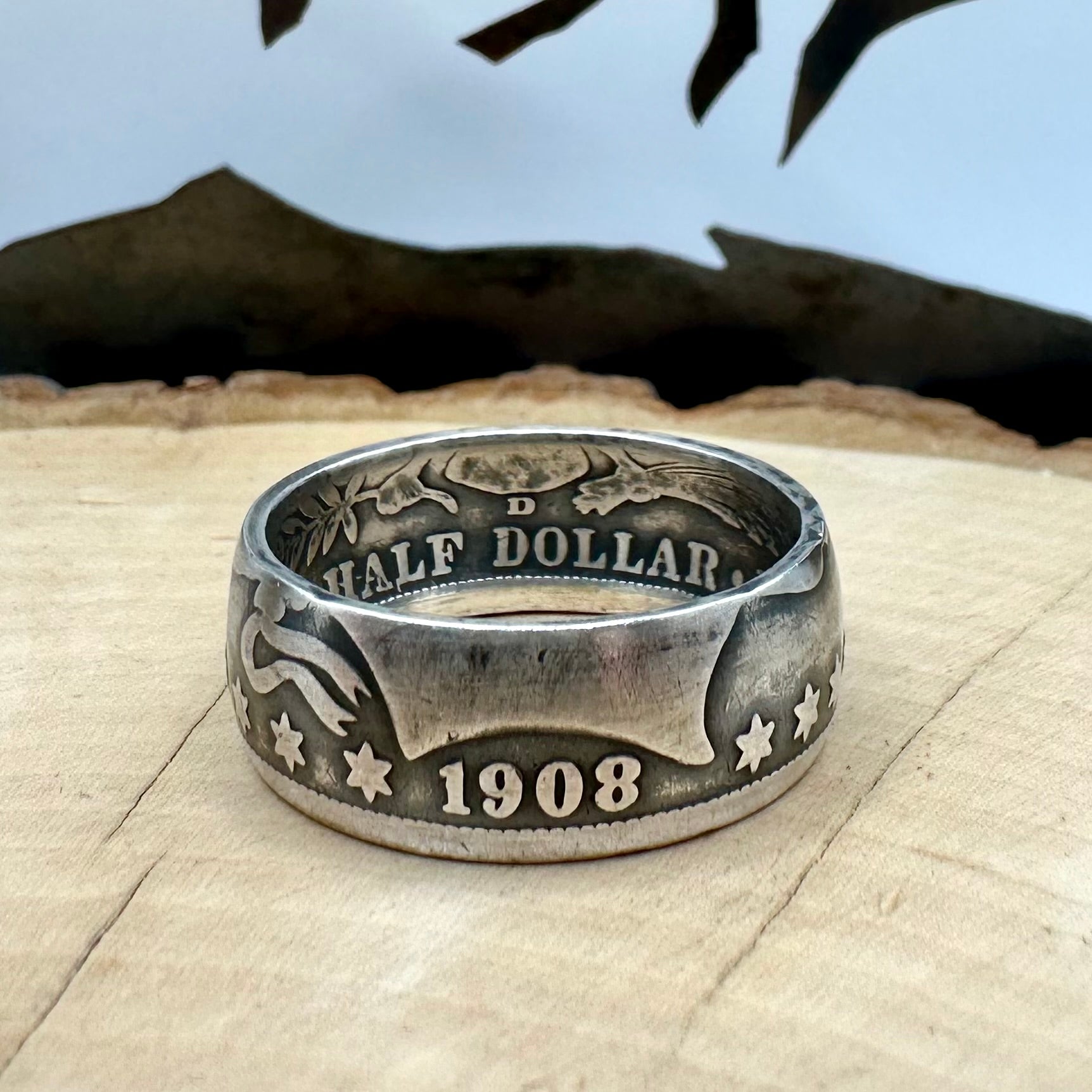 U.S. Barber Silver Half Dollar Coin Ring (front) on a light wood round with metal mountains in the background. - Mountainstruck Coin Jewelry