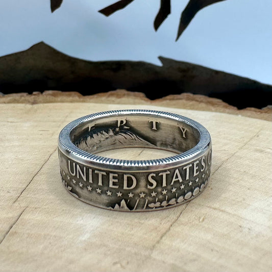 U.S. JFK Silver Half Dollar Coin Ring (reverse) on a light wood round with a metal mountain background. - Mountainstruck Coin Jewelry