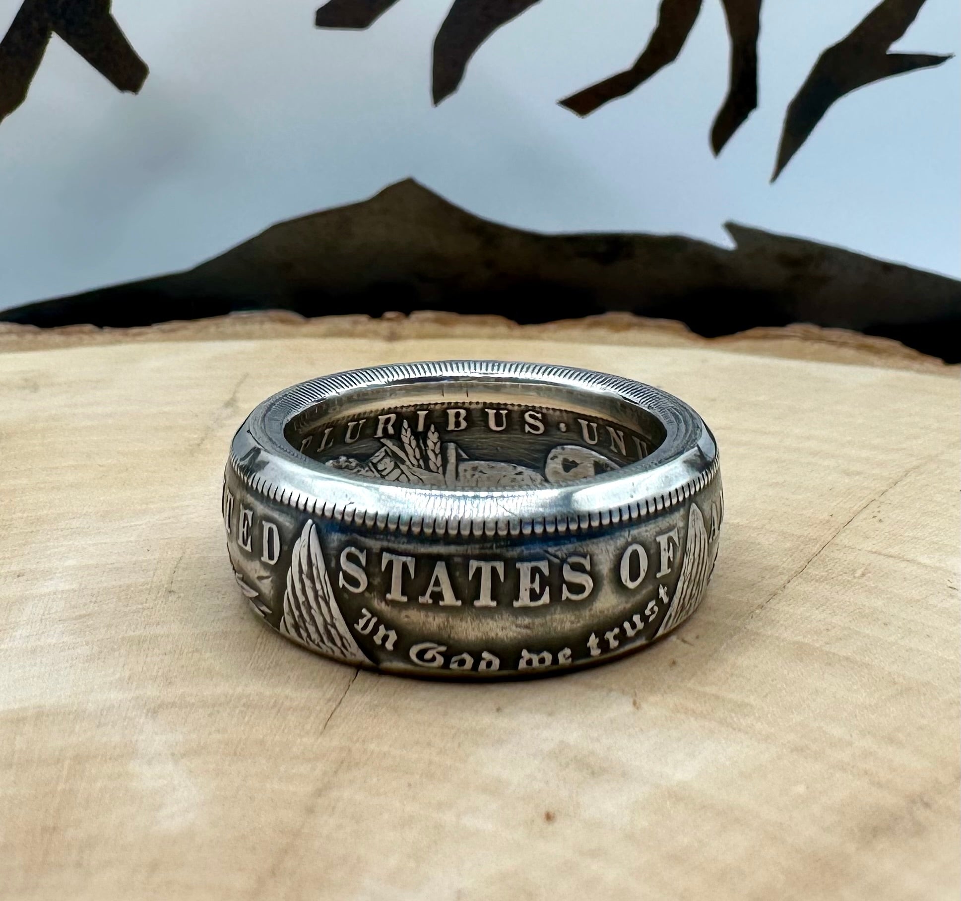 U.S. Morgan Dollar Silver Coin Ring patina on a light wood round with a metal mountain background. - Mountainstruck Coin Jewelry