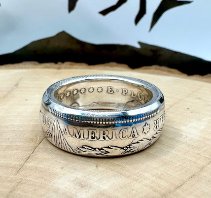 U.S. Morgan Dollar Silver Coin Ring shine on a light wood round with a metal mountain background. - Mountainstruck Coin Jewelry