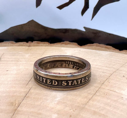 U.S. Presidential Dollar Coin Ring (Reverse) patina on a light wood round with a metal mountain background. - Mountainstruck Coin Jewelry