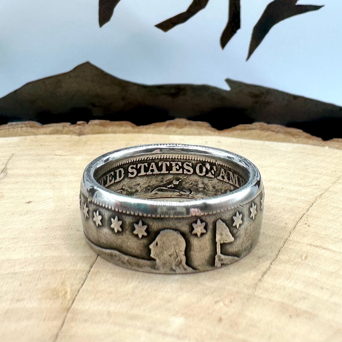 U.S. Seated Liberty Silver Half Dollar Coin Ring (patina) on a light wood round with a metal mountain background. - Mountainstruck Coin Jewelry