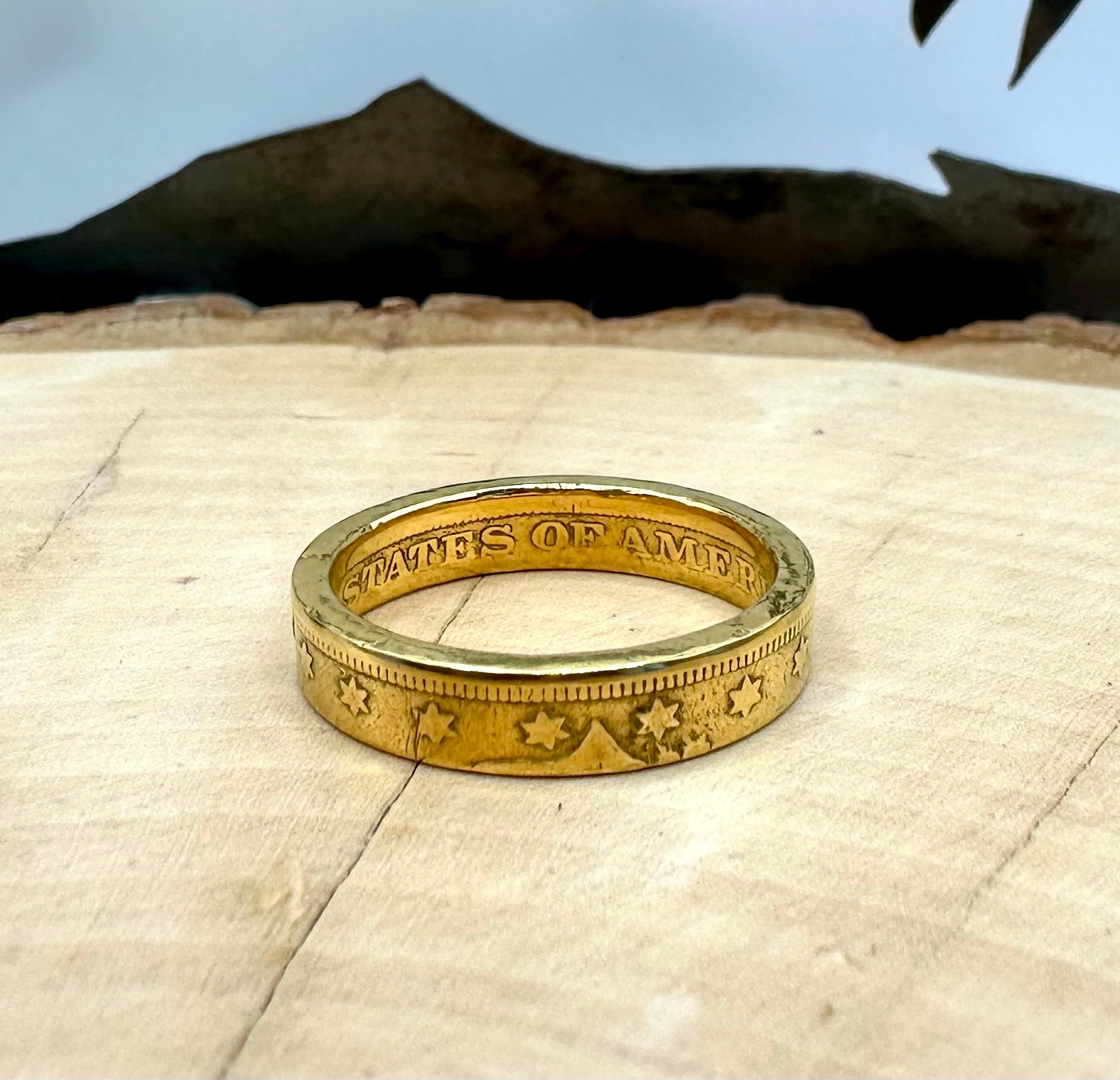 "V" Nickel Coin Ring 24k gold plated on a light wood round with a metal mountain background. - Mountainstruck Coin Jewelry