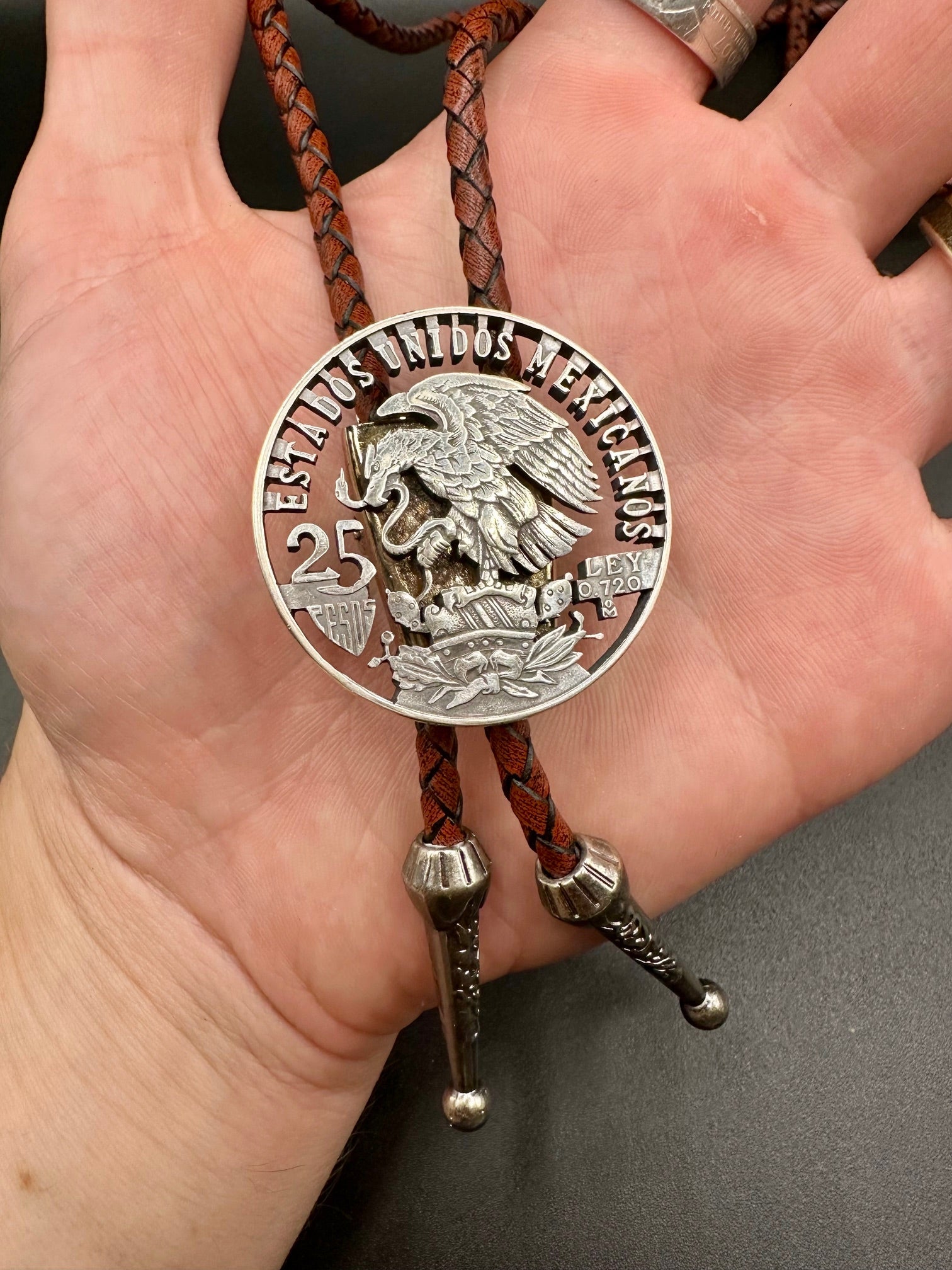 Mexico Silver 25 Pesos Hand Cut Coin Bolo Tie with darker brown leather cord on a hand for display.