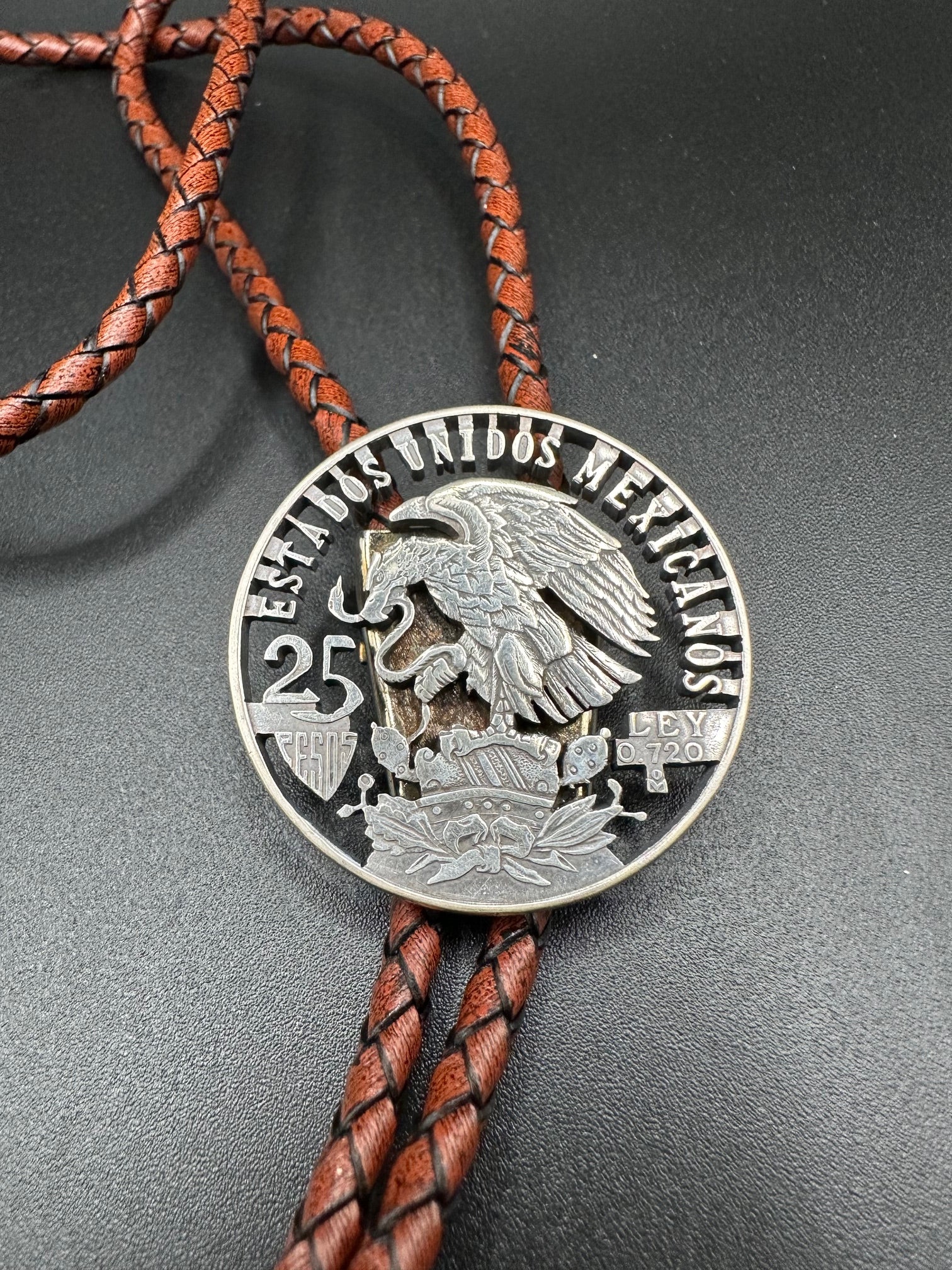 Mexico Silver 25 Pesos Hand Cut Coin Bolo Tie with darker brown leather cord on black background.