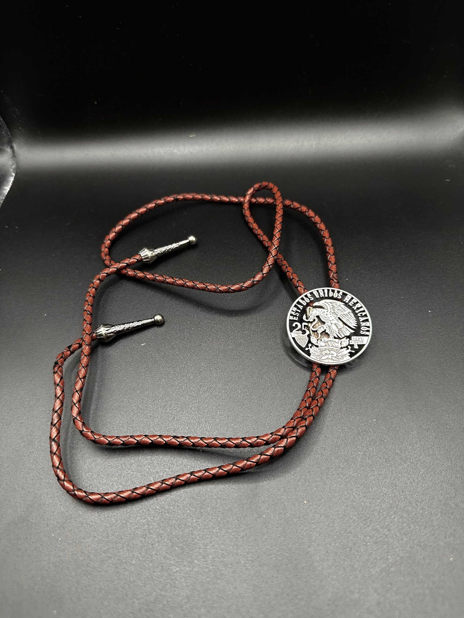 Mexico Silver 25 Pesos Hand Cut Coin Bolo Tie with darker brown leather cord on black background.