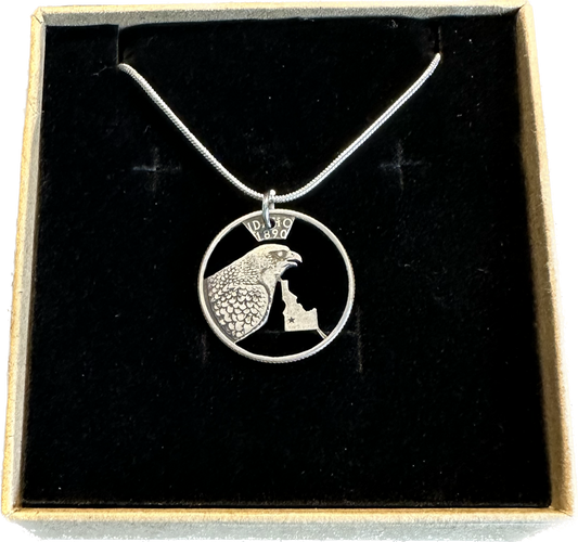 Idaho State Hand Cut Quarter Necklace with a snake chain in a brown kraft box on a black velvet background.
