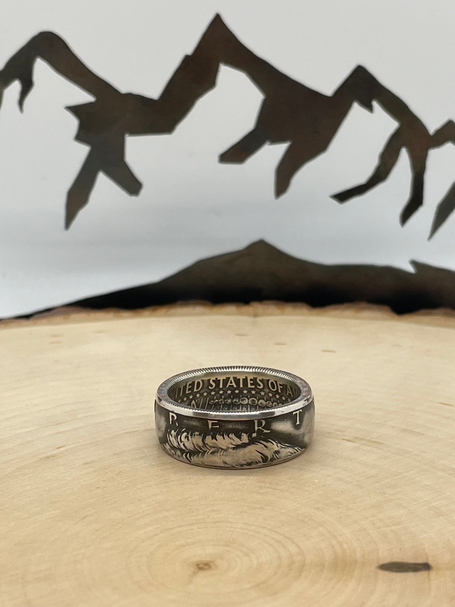 JFK Silver Half Dollar Coin Ring (front) on a light wood round with mountain outline in background.