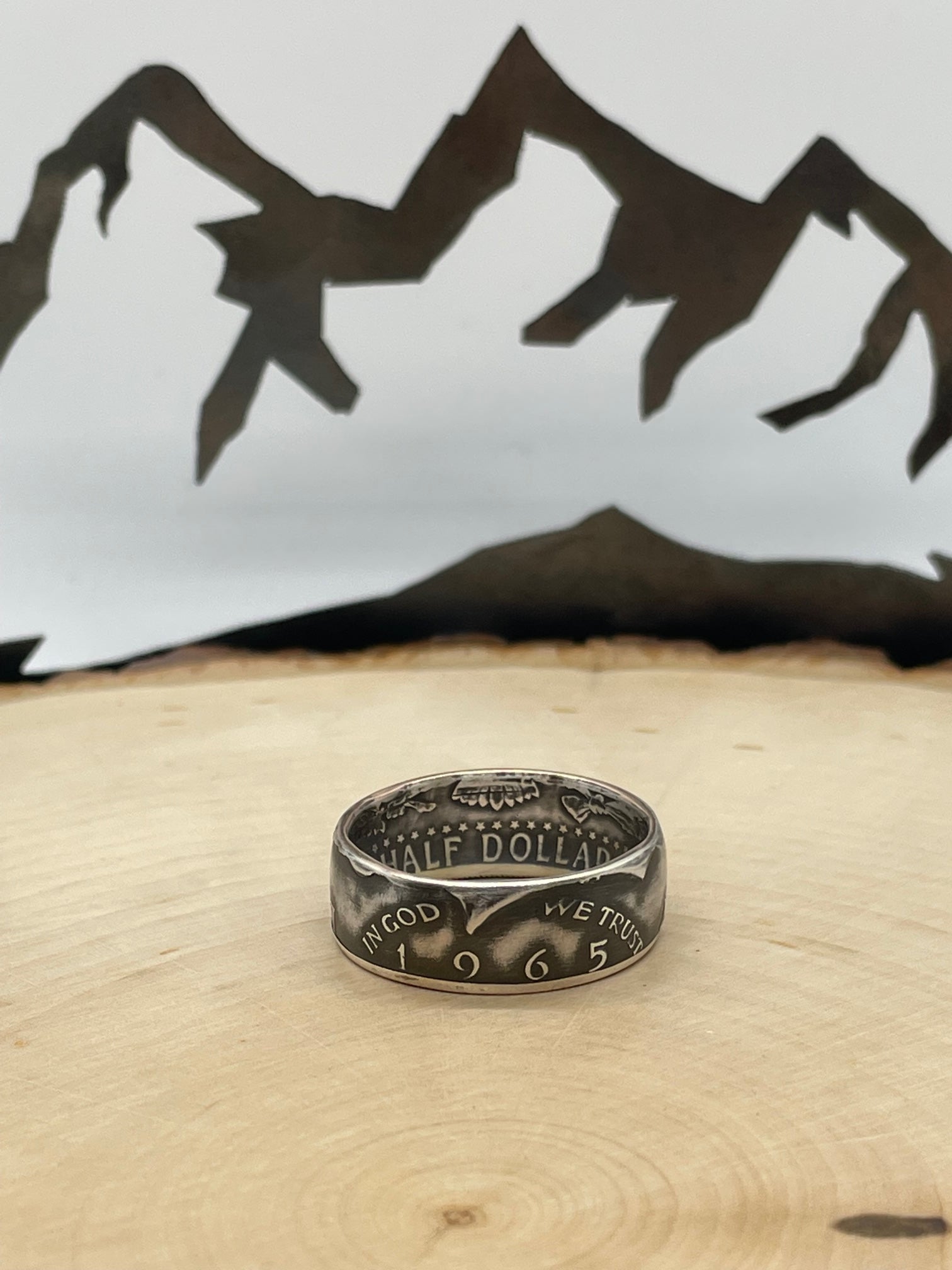 JFK Silver Half Dollar Coin Ring (front) on a light wood round with mountain outline in background.