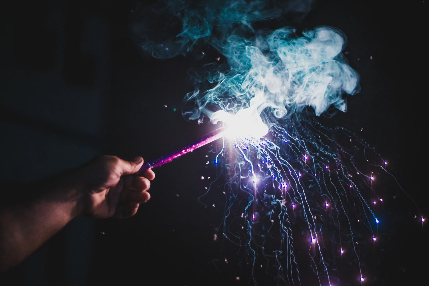 Hand holding lit firework with smoke and purple sparks in front of a black background. -Mountainstruck Coin Jewelry
