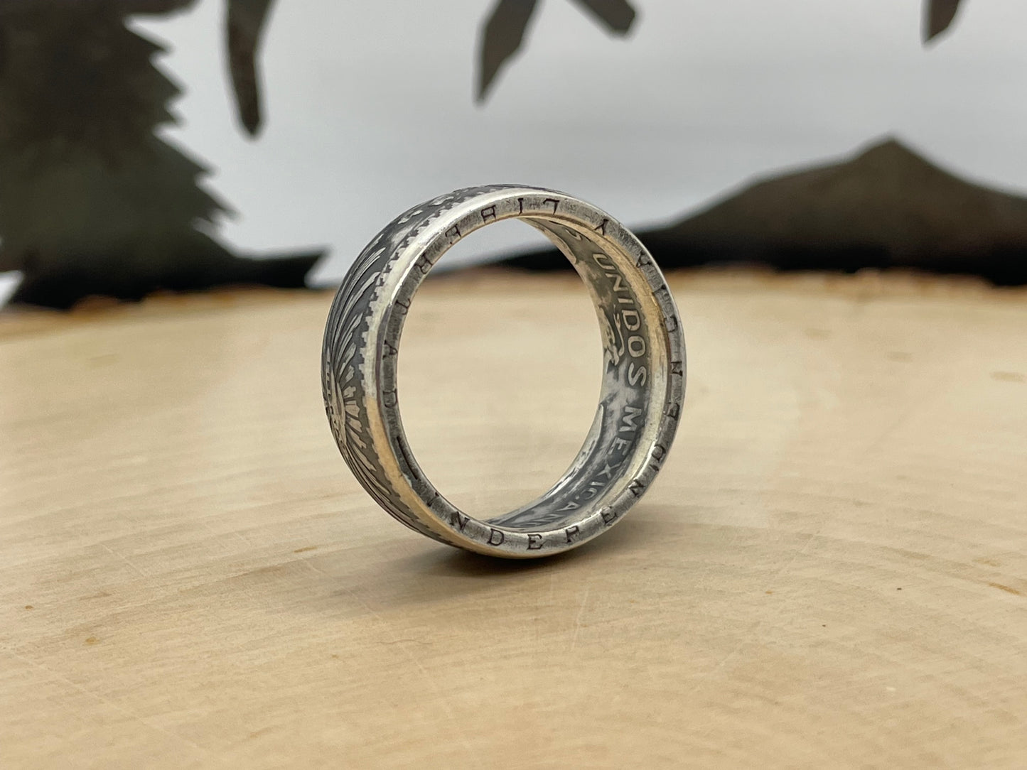 Mexico 50 Centavos Silver Coin Ring (Rare) on its edge on a light wood round with mountain outline in the background. - Mountainstruck Coin Jewelry