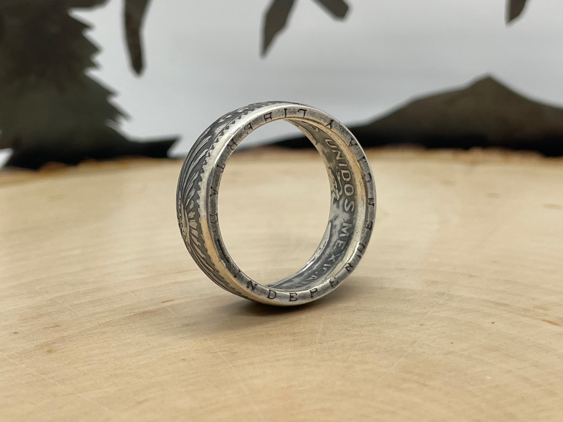 Mexico 50 Centavos Silver Coin Ring (Rare) on its edge on a light wood round with mountain outline in the background. - Mountainstruck Coin Jewelry