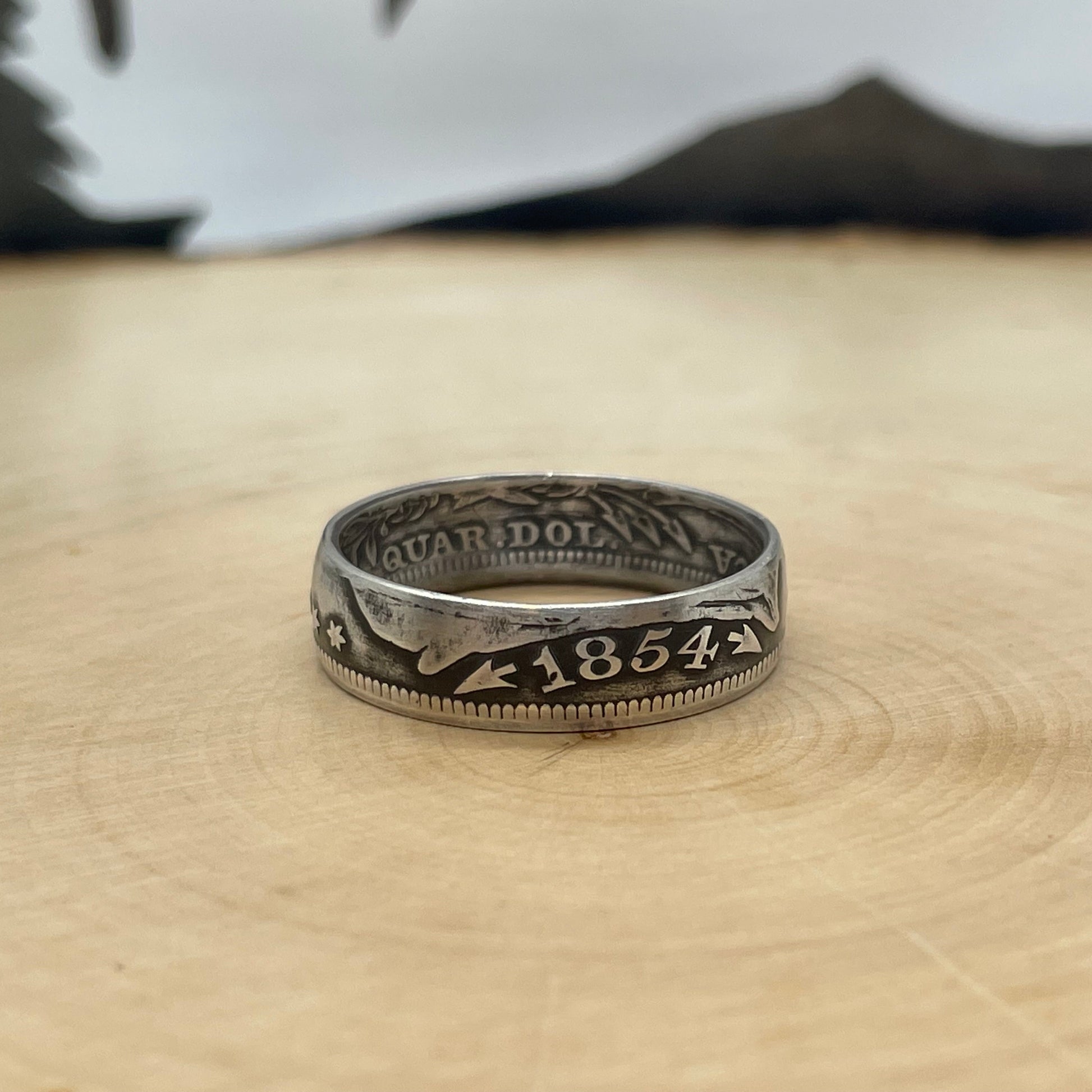 U.S. Seated Liberty Silver Quarter Coin Ring on a light wood round with metal mountain background. - Mountainstruck Coin Jewelry