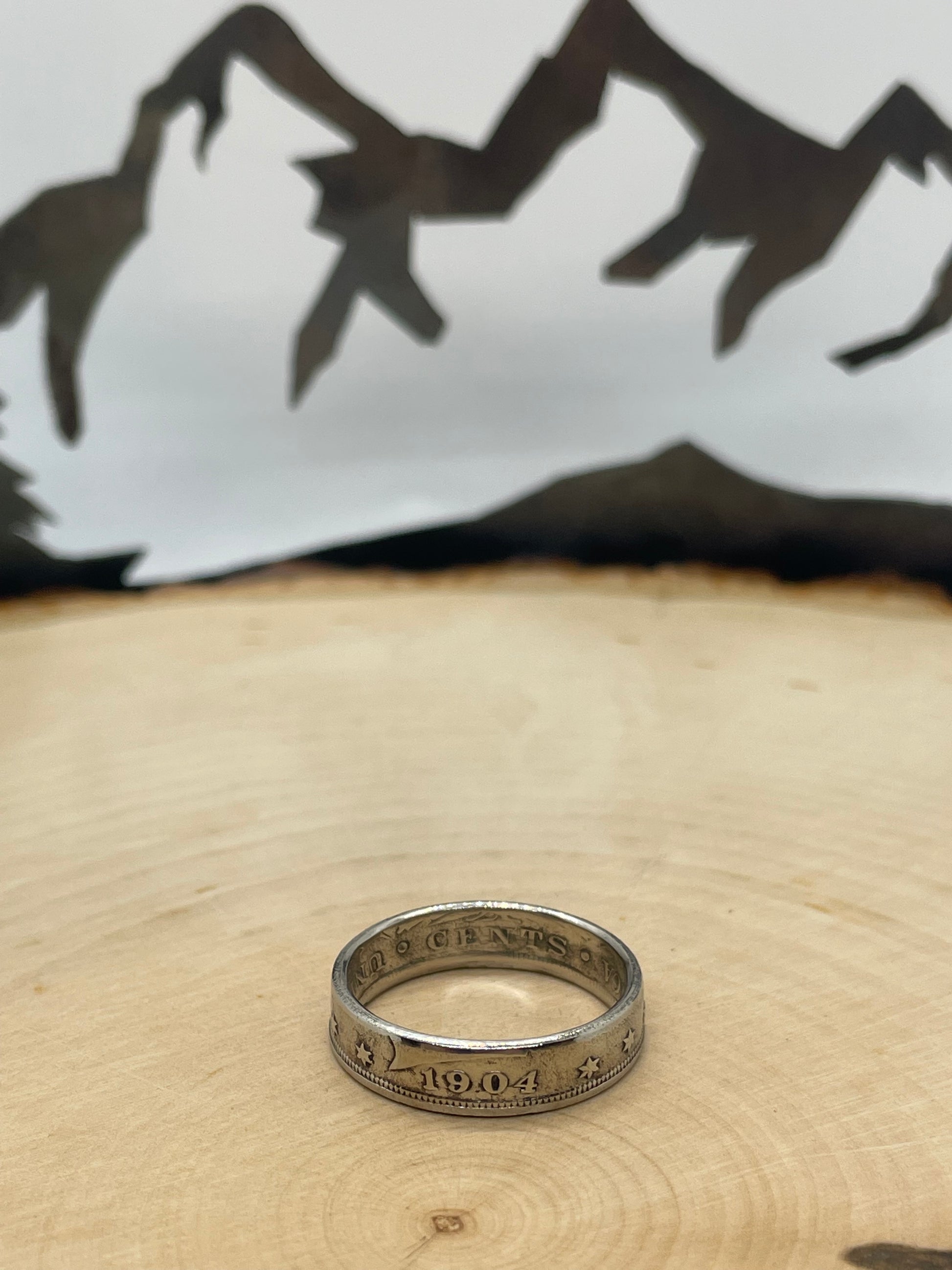 "V" Nickel Coin Ring on wood round with mountain outline in background.
